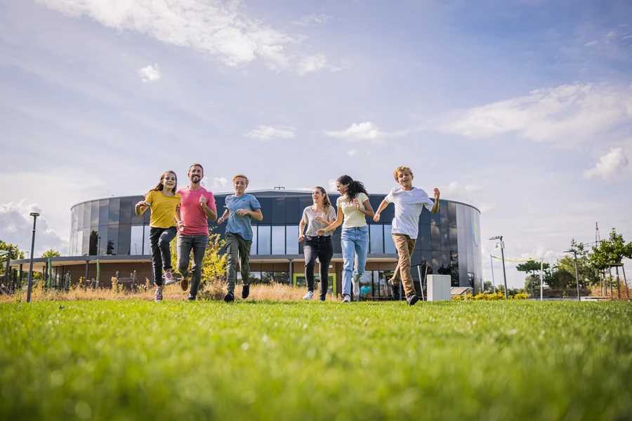 Familie l&auml;uft auf der Wiese im Themenpark, im Hintergrund ist das Geb&auml;ude der KLIMA ARENA zu sehen.