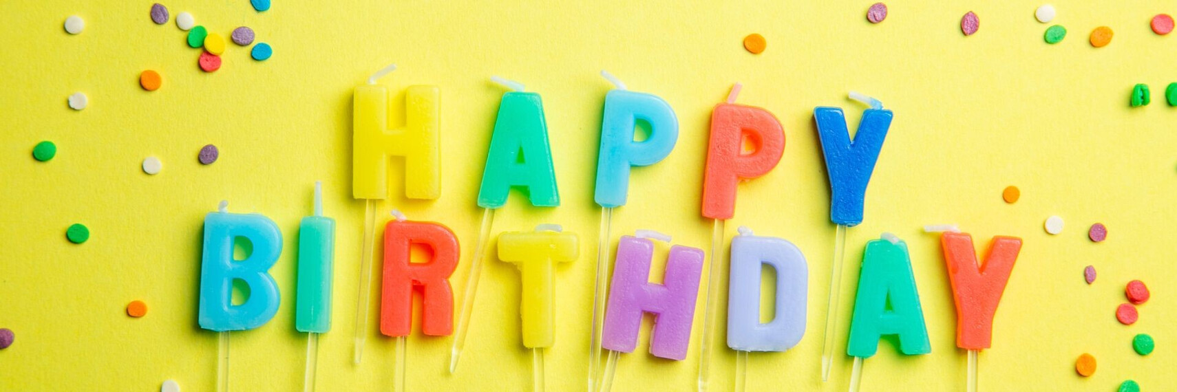 Schriftzug Happy Birthday aus Kerzen vor einem gelben Hintergrund mit Konfetti.
