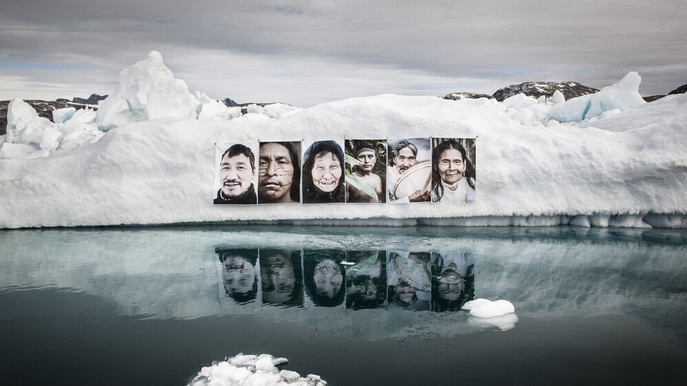 Gro&szlig;e Portr&auml;ts von Personen aus indigenen V&ouml;lkern weltweit h&auml;ngenan einem Eisberg.