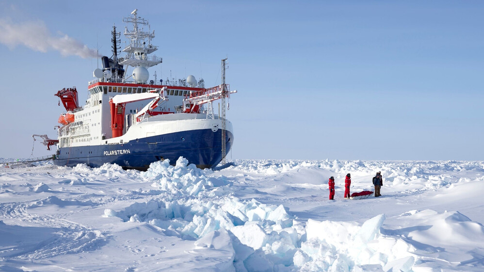 Der Eisbrecher Polarstern im arktischen Eis und drei Wissenschaftler stehen in Eisfeld davor.