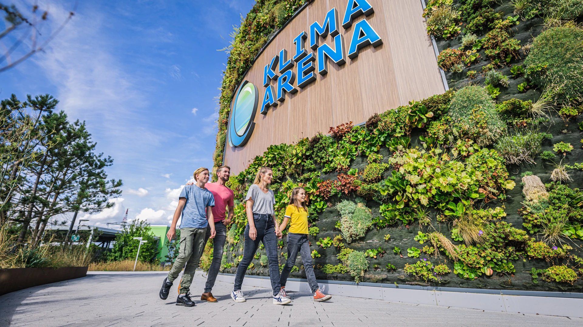 Familie l&auml;uft auf dem Vorplatz der KLIMA ARENA entlang der begr&uuml;nten Fassade mit Logo.