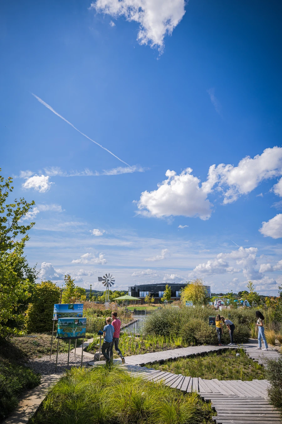 Blick vom angelegten Moor &uuml;ber den Themenpark auf das Geb&auml;ude der KLIMA ARENA in der Ferne. Besuchende auf Holzstegen &uuml;ber dem Moor.