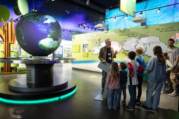Eine Gruppe von Kindern und Erwachsenen nimmt in einem ineraktiven Ausstellungsst&uuml;ck an einer F&uuml;hrung teil.